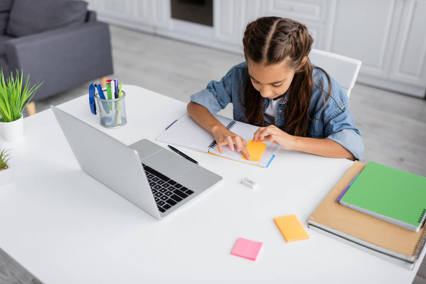 Κορίτσι εφαρμογή κολλώδη σημείωμα στο σημειωματάριο κοντά γραφική ύλη και φορητό υπολογιστή στο σπίτι  - Φωτογραφία, εικόνα