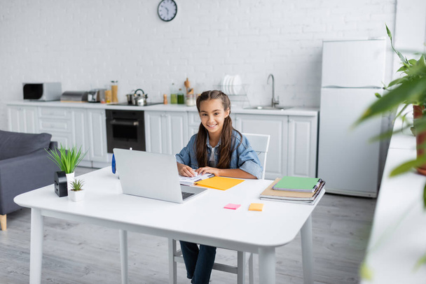 Preteen kid smiling at camera near notebooks and laptop in kitchen  - Photo, Image