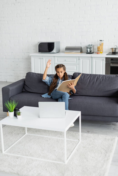 Preteen kid with notebook raising hand near laptop on couch at home  - Valokuva, kuva