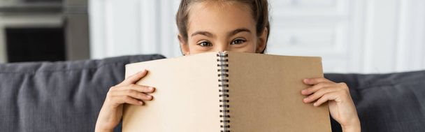 Preteen kid covering face with notebook on couch at home, banner  - Fotoğraf, Görsel