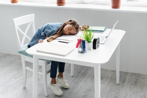 Preteen girl sleeping near stationery and laptop on table at home  - Foto, Bild