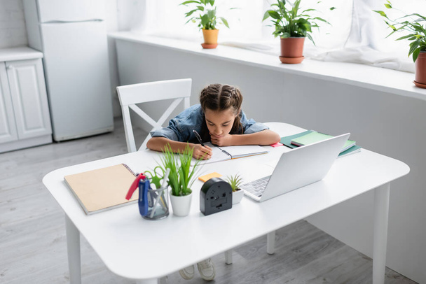 Child writing on notebook near laptop and plants at home  - Φωτογραφία, εικόνα