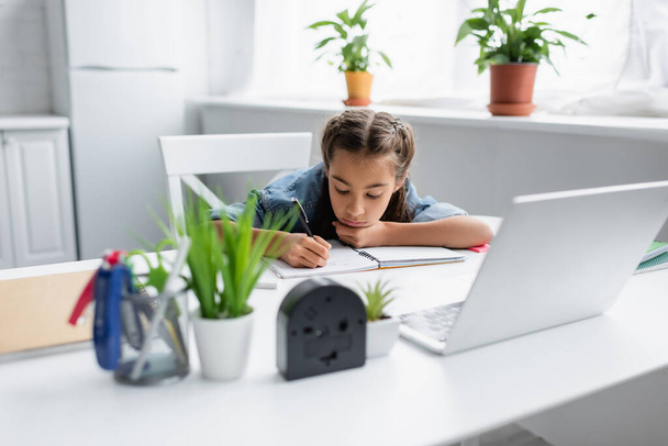 Gyermek írás a másolókönyv közelében elmosódott laptop és a növények otthon  - Fotó, kép