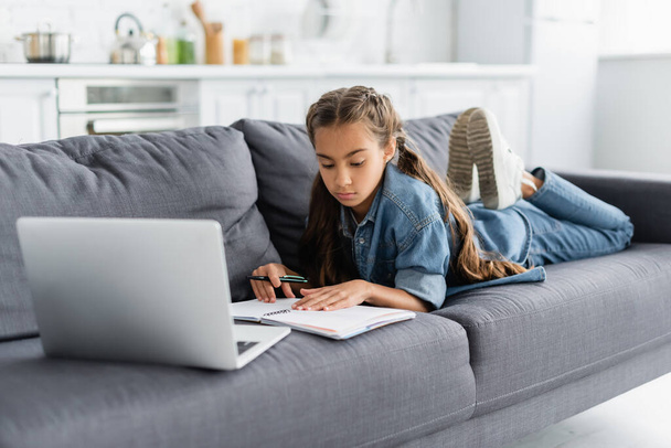 Kid holding pen near notebook and blurred laptop on couch  - Foto, Imagem