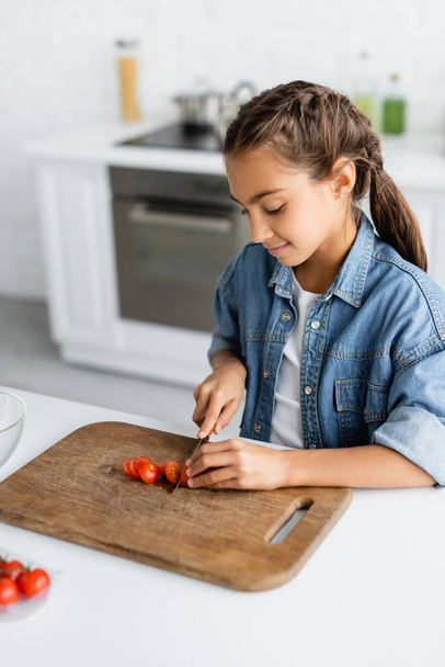 Усміхнений дитина ріже вишневий помідор на дошці для різання деревини на кухні
  - Фото, зображення