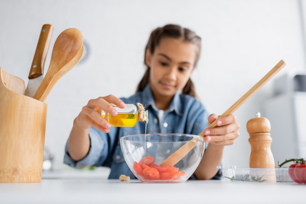 Menina borrada derramando azeite na tigela com tomates cereja na cozinha  - Foto, Imagem