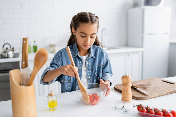 Criança pré-adolescente misturando tomates cereja em tigela perto de tábua de corte e azeite na cozinha  - Foto, Imagem