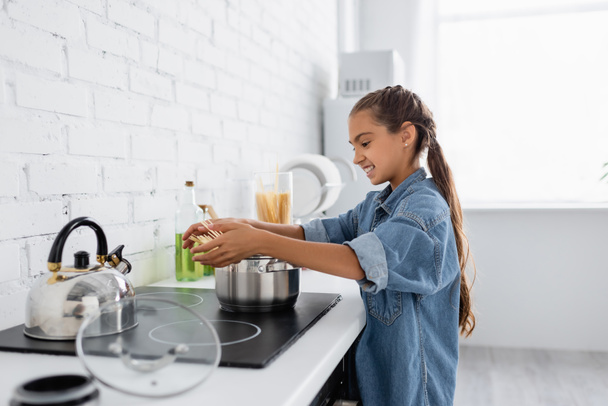 Vista lateral de niña alegre sosteniendo macarrones cerca de la cacerola y hervidor de agua en la cocina  - Foto, Imagen