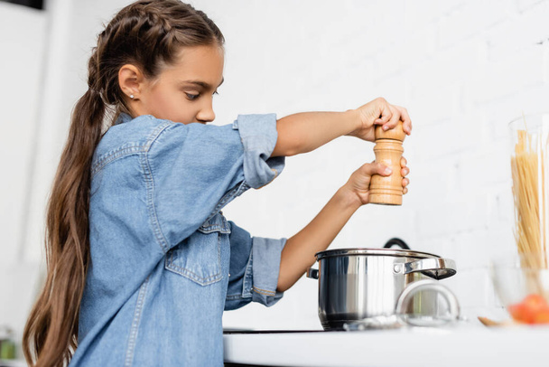 Preteen kid holding salt mill near saucepan in kitchen  - Fotó, kép