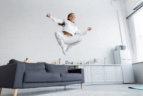 Low angle view of cheerful girl in sportswear jumping near couch in kitchen  - Photo, Image