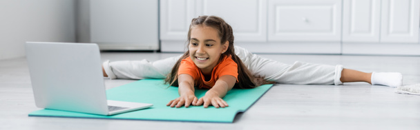 Улыбающийся ребенок делает раскол возле ноутбука на тренажерном коврике дома, баннер  - Фото, изображение