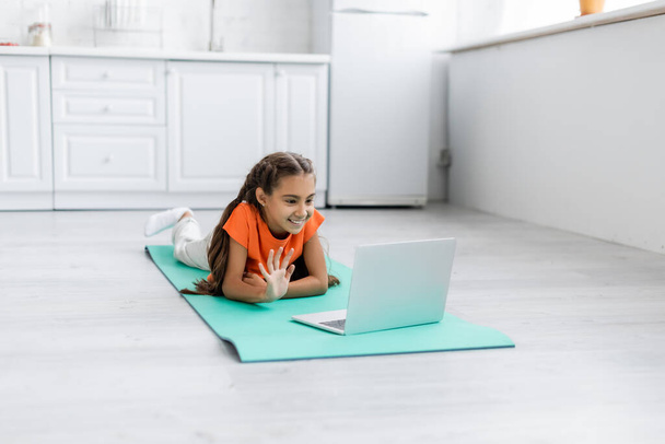 Smiling preteen girl having video call on laptop on fitness mat at home  - Foto, imagen