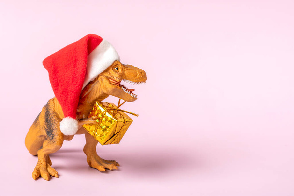 Dinosaur Rex in red Santa Claus hat holds golden gift box in its paws on pink background New Years Eve or Christmas Eve Art holiday card Creative idea for Merry xmas concept. - Photo, Image
