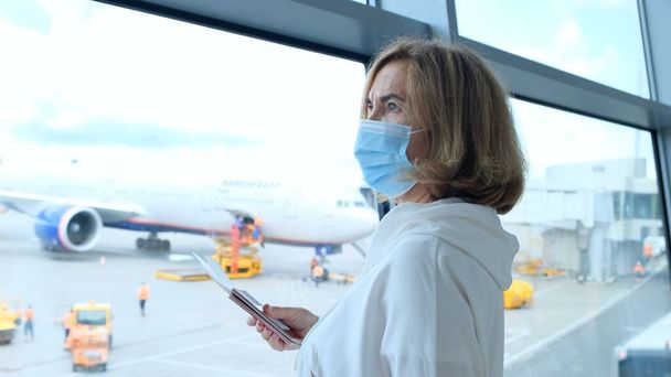 ενήλικη γυναίκα που φοράει προστατευτική μάσκα στέκεται στο παράθυρο του τερματικού σταθμού του αεροδρομίου περιμένοντας την αναχώρηση πτήσης λόγω ταξιδιωτικών περιορισμών λόγω της πανδημίας του κορωναϊού, ηλικιωμένης ηλικίας 50 ετών - Φωτογραφία, εικόνα