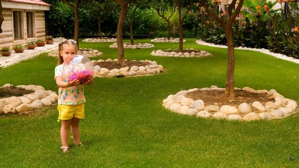 Милая маленькая девочка, 3 года, с двумя хвостиками на голове, в разноцветной футболке и желтых шортах, играет с разноцветным мячом на зеленом лугу в саду своего дома. При этом - Фото, изображение