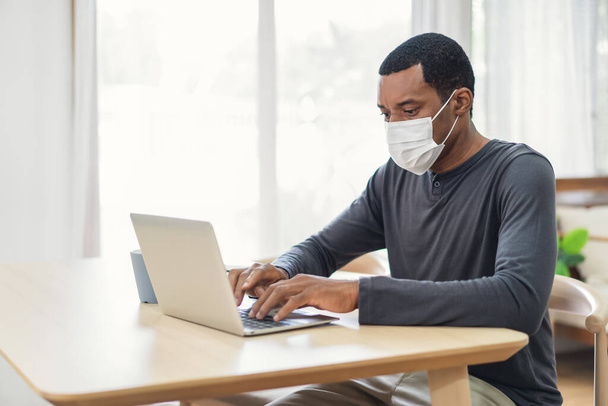 Siyah iş adamı evdeki bir masada dizüstü bilgisayarla otururken tıbbi koruyucu maske takıyor. Kendini izole etme karantinası, yeni normal. Coronavirus Covid-19 Salgını sırasında Sosyal Mesafe - Fotoğraf, Görsel