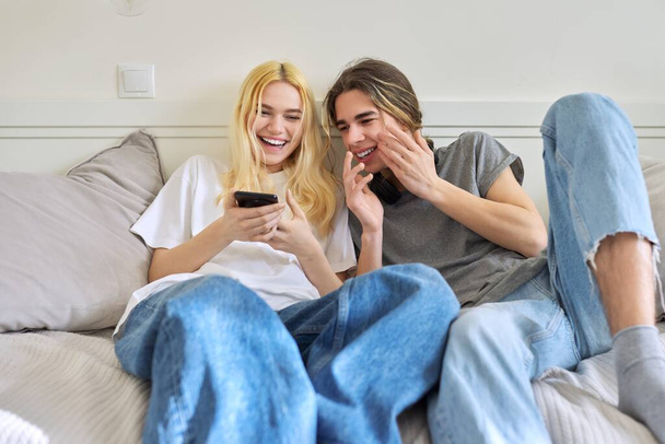 Couple of teenagers having fun together, sitting on the couch looking into the smartphone - Fotografie, Obrázek