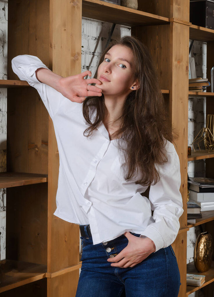 Brunette caucasian girl stands near bookshelf in white blouse and blue jeans - Fotoğraf, Görsel