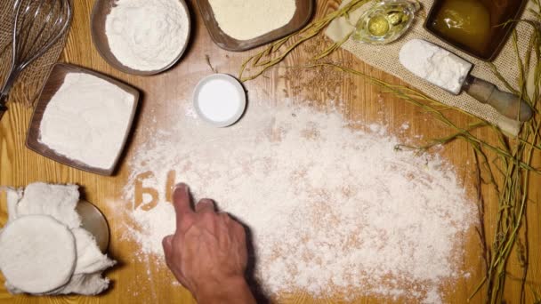 Pan casero. La mano de un hombre escribe con su dedo sobre la harina "pan sin levadura" en ruso.En la mesa están los ingredientes para hacer pan de centeno: harina (centeno, trigo, maíz) masa madre, sal, miel, aceite.. - Metraje, vídeo