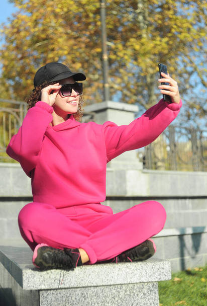Mujer sosteniendo el teléfono móvil en las manos y haciendo selfies al aire libre. Cabello rizado mujer joven en ropa deportiva magenta fuera - Foto, imagen