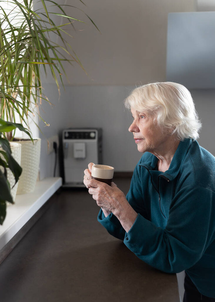 Een aantrekkelijke grijs-harige 85-jarige oudere vrouw drinkt espresso in de keuken en kijkt uit het raam. - Foto, afbeelding