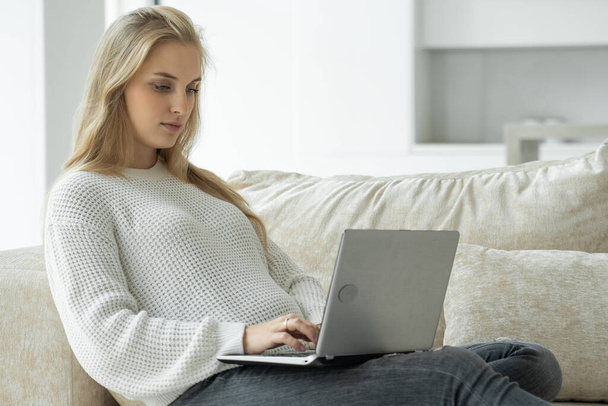 Pregnant woman is sitting on a beige sofa with a laptop. The concept of a pregnant woman working at home - 写真・画像