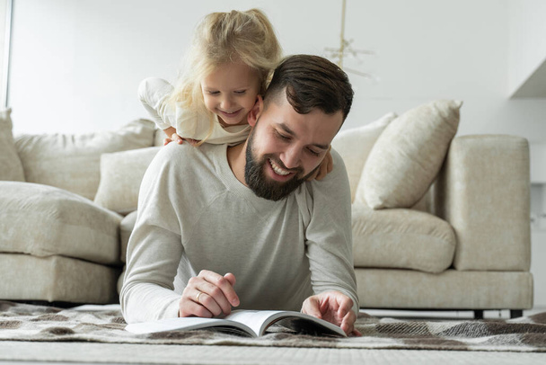 Bearded father reads a fairy tale for his little daughter lying on the floor of the house. Happy family lying on the living room floor - 写真・画像