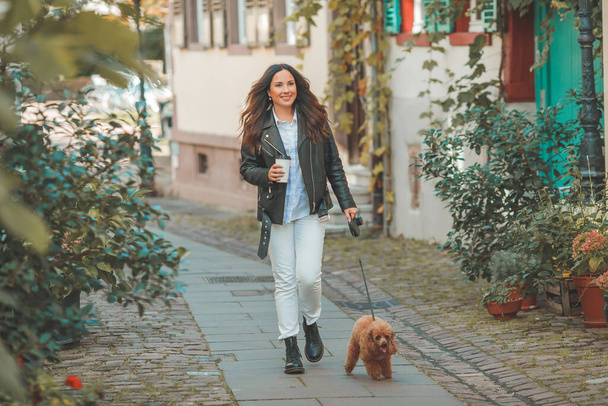 A young beautiful woman with her poodle dog is walking in a European city. Happiness. Little pet. Fall. - Photo, Image