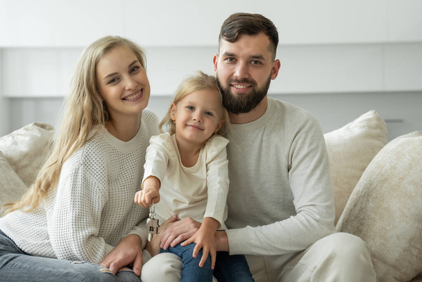 Little girl holds the keys to a new family home in her hands. Portrait of a smiling young married couple and a cute girl showing the keys - Fotoğraf, Görsel