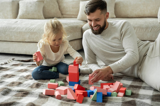 Bearded father and daughter play dice together at home, sitting on the floor in the living room. - 写真・画像