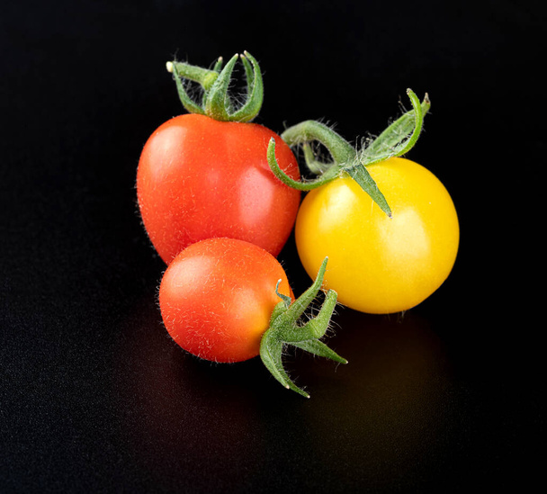 Młody, świeży, piękny, żółty pomidor wiśniowy i dwa bardzo małe, dojrzałe, czerwone pomidory wiśniowe na błyszczącym, czarnym tle z odbiciem. Zbliżenie - Zdjęcie, obraz
