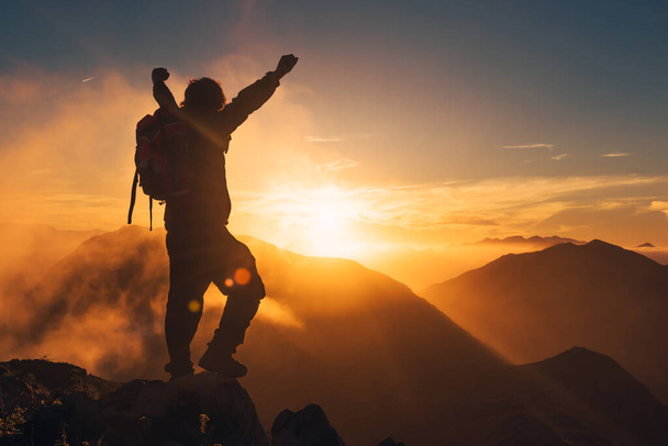 verlicht silhouet van wandelaar met rugzak die zijn armen opsteekt in overwinningstekens terwijl hij naar de zonsondergang kijkt. bergsport concept. - Foto, afbeelding