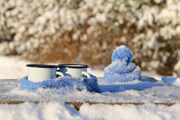 Dva bílé železné hrnky čaje zabalené do šály vedle modrého pleteného klobouku stojí na dřevěném stole na pozadí zimní krajiny. - Fotografie, Obrázek