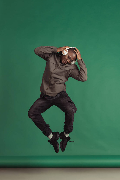 Studio shot of african man in headphones jumping and listening music isolated over dark green color background - Fotografie, Obrázek