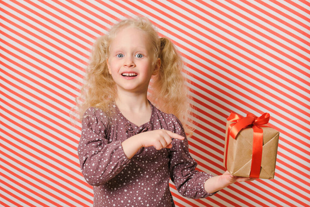 cheerful preschooler girl with a gift on a red striped background, a gift with a red ribbon in the hands of a girl with blond hair, boxing day, gifts for birthday and christmas, holiday gifts - Photo, Image