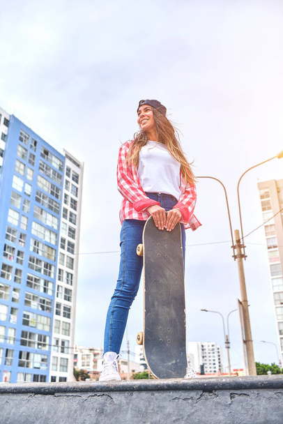 Όμορφη νεαρή γυναίκα με καπέλο κάθεται στο skatepark στη ράμπα με το longboard της. - Φωτογραφία, εικόνα