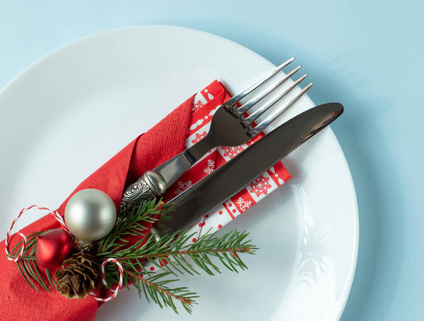 Příbory zabalené v červeném ubrousku s vánoční výzdobou je na bílém talíři na modrém pozadí. Pohled z horního bodu. - Fotografie, Obrázek