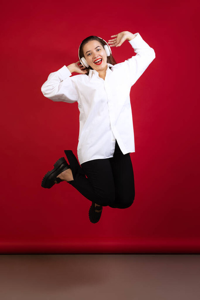 Optimistic young beautiful girl wearing white shirt laughing and jumping isolated on dark red studio background. Concept of emotions, fashion - Foto, Imagen