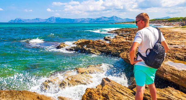 Male traveler and tourist is watching the rough natural coastal and beach landscape panorama with turquoise water waves mountains rocks boulders in Can Picafort on Balearic island Mallorca in Spain. - 写真・画像