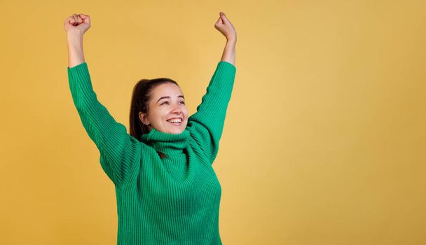 Dynamic image of young cute girl, student wearing green sweater raises hands isolated on yellow studio background. Concept of emotions, fashion - Foto, afbeelding