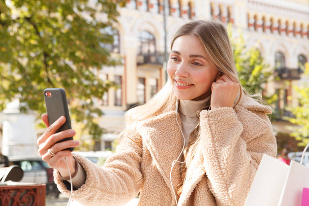 Lovely woman smiling, putting in her earphones, usign smart phone outdoors - Photo, Image
