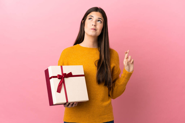 Teenager Brazilian girl holding a gift over isolated pink background with fingers crossing and wishing the best - Foto, Bild