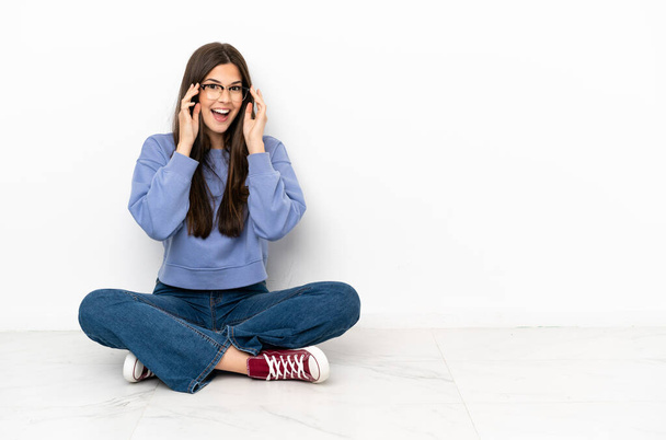Jonge vrouw zittend op de vloer met een bril en verrast - Foto, afbeelding