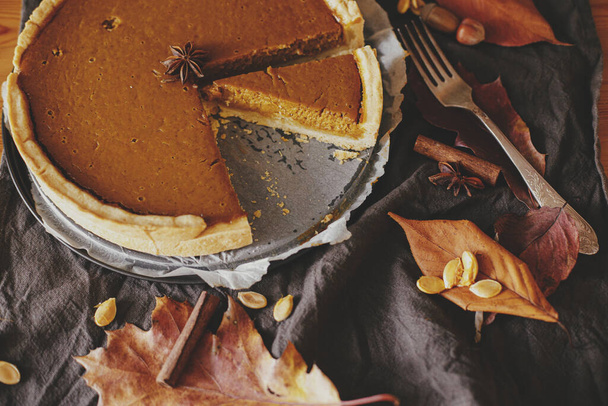 Happy Thanksgiving. Pumpkin pie on parchment paper on rustic table with linen napkin, autumn leaves, anise, cinnamon and cutlery. Homemade pumpkin tart recipe. Top view - Photo, Image
