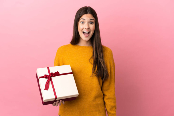 Teenager Brazilian girl holding a gift over isolated pink background with surprise facial expression - Photo, Image