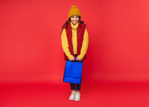 happy kid in winter hat hold shopping bags on red background, shopping - Valokuva, kuva