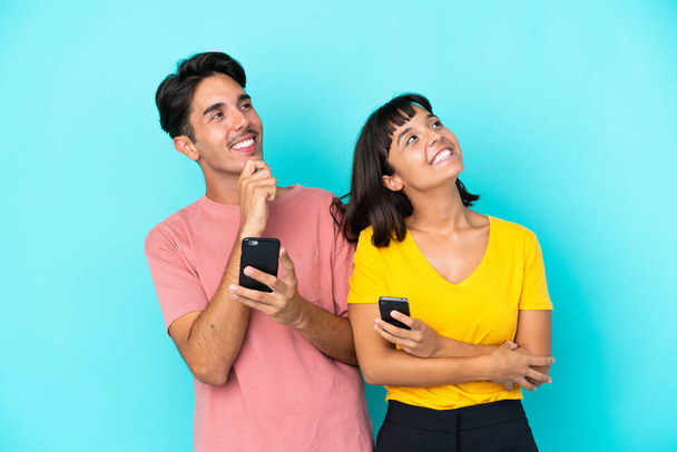Young mixed race couple holding mobile phone isolated on blue background looking up while smiling - 写真・画像