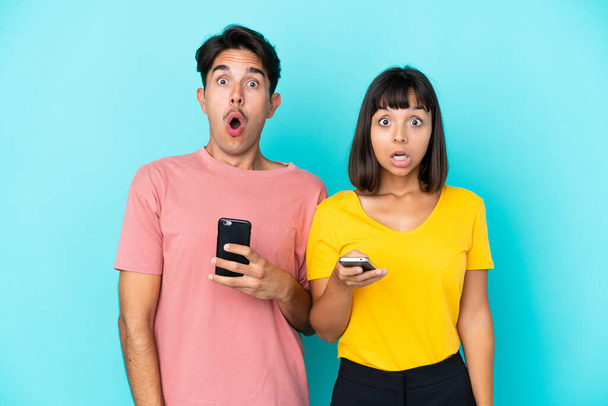 Молодая пара смешанной расы держит мобильный телефон на синем фоне с удивлением и шокированным выражением лица - Фото, изображение