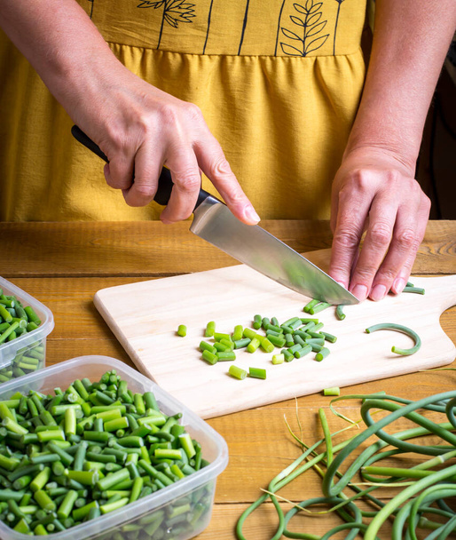 Les flèches d'ail vert sont coupées avec un couteau sur une planche en bois. Préparation des approvisionnements alimentaires pour l'hiver. - Photo, image