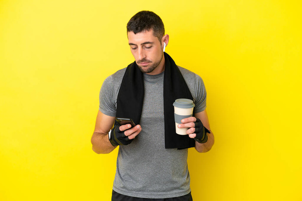 Молодий спортивний кавказький чоловік ізольований на жовтому фоні, тримаючи каву, щоб забрати і мобільний
 - Фото, зображення